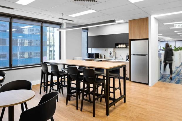 Contemporary commercial office breakout kitchen and dining area with a large timber island table surrounded by black bar stools and café tables. The space features light timber flooring, black cabinetry and splashback accents, stainless-steel appliances, expansive windows, and bright linear ceiling lighting, with an open-plan office visible beyond.