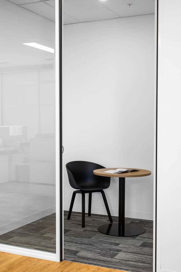 A minimalist office breakout or waiting area enclosed by clear glass partitions, with a black molded chair and small round timber-topped pedestal table holding a magazine. The space has white walls, a suspended white ceiling, dark patterned carpet tiles, and timber flooring visible outside the glass enclosure.