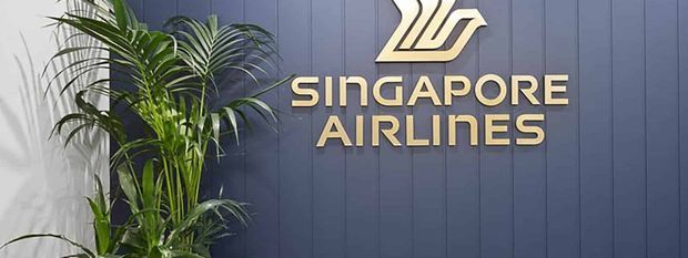 A Singapore Airlines logo and large gold lettering mounted on a dark blue vertically panelled wall, with a tall indoor palm plant positioned to the left. The image appears to show a branded commercial or corporate interior feature wall.