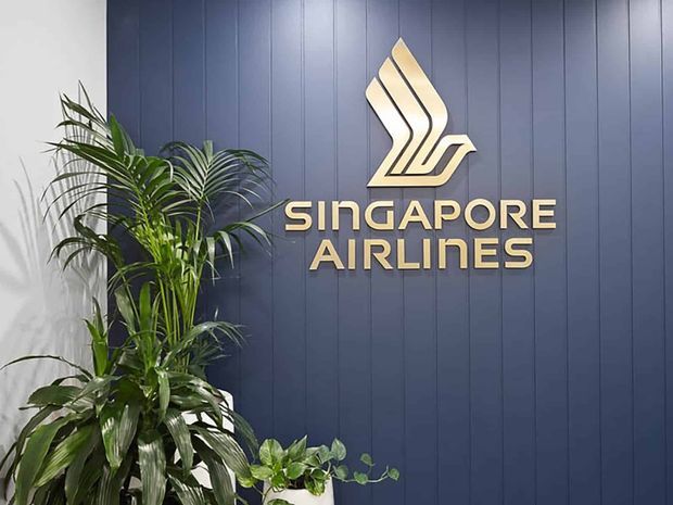 A branded reception or office feature wall for Singapore Airlines, with raised gold lettering and logo mounted on dark navy vertical wall panelling. Large leafy indoor plants in white planters sit in the foreground.