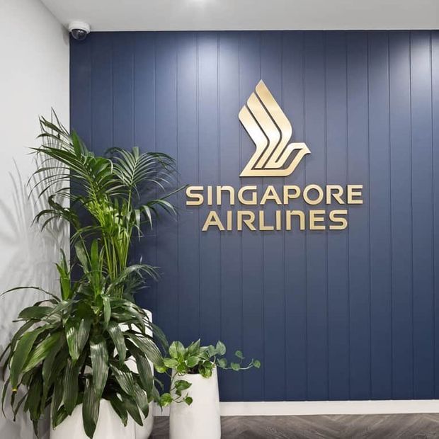 A branded reception or entry area featuring a large gold Singapore Airlines logo and lettering mounted on a dark navy vertically panelled feature wall. Lush potted tropical plants sit in white planters in front of the wall, with a white ceiling and dark timber-look flooring.