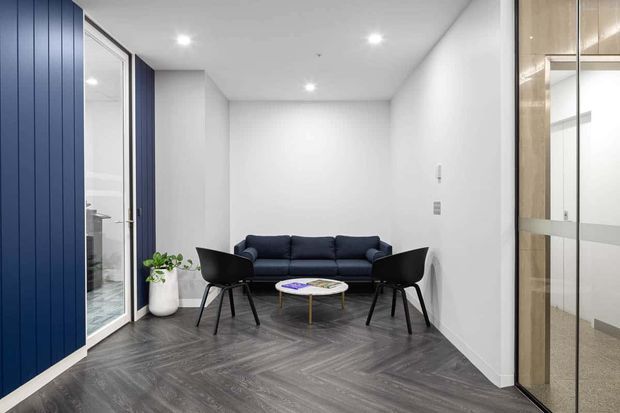 A compact, contemporary waiting or reception area with a dark navy sofa, two black lounge chairs, and a small round coffee table. The space has white walls and ceiling with recessed downlights, dark grey herringbone-pattern flooring, a deep blue vertical-panel feature wall, glazed doors, and a reflective glass partition.