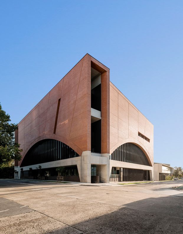 Exterior architectural photograph of a large contemporary institutional or commercial building on a street corner. The building has a strongly geometric, monolithic form clad in warm reddish-brown panels, with two prominent tall arched openings filled with dark vertical louvres and recessed balconies or circulation spaces. A pale concrete base, glazed ground-level frontage, and a narrow vertical slot opening define the facade. The image is taken in clear daylight from across a broad paved street or forecourt.