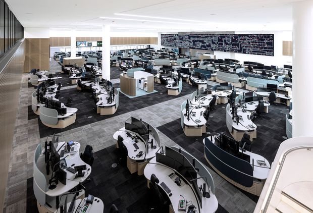 A large, open-plan commercial operations centre or control room viewed from an elevated position. Numerous curved workstation clusters with computer monitors, phones and office equipment are arranged across a broad floor, with partial-height acoustic screens and a large wall-mounted information display. The space has a bright white ceiling, dark patterned carpet tiles, light grey flooring sections, pale wood-toned partitions and extensive glazing or reflective panels along the left wall. The overall design is highly organised, modern and functional.
