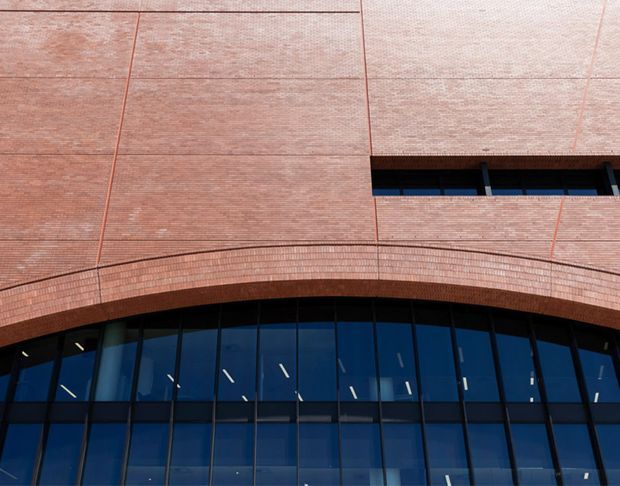 Close-up upward view of a contemporary commercial building facade, combining extensive reddish brick cladding with large dark-framed blue reflective glazing. A curved projecting brick band arches above the glazed lower facade, while narrow horizontal window openings punctuate the upper wall.