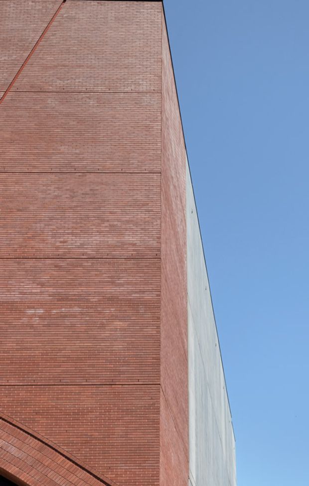 Upward-looking architectural exterior photograph of a tall building corner. The dominant facade is composed of fine red brickwork with horizontal coursing and subtle recessed bands, meeting a pale smooth side wall at a sharp vertical edge. A clear blue sky occupies the right side of the frame. No windows, doors, interiors, or landscaping are visible.