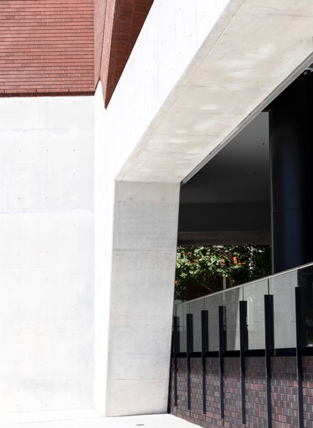 Close-up exterior architectural view showing a contemporary concrete entry or covered threshold. Pale raw concrete walls and a large angled concrete beam frame a dark recessed opening, with a glass balustrade supported by black vertical posts and a low red-brown brick wall. Red brick cladding is visible at the upper left, and greenery appears through the opening in the background.