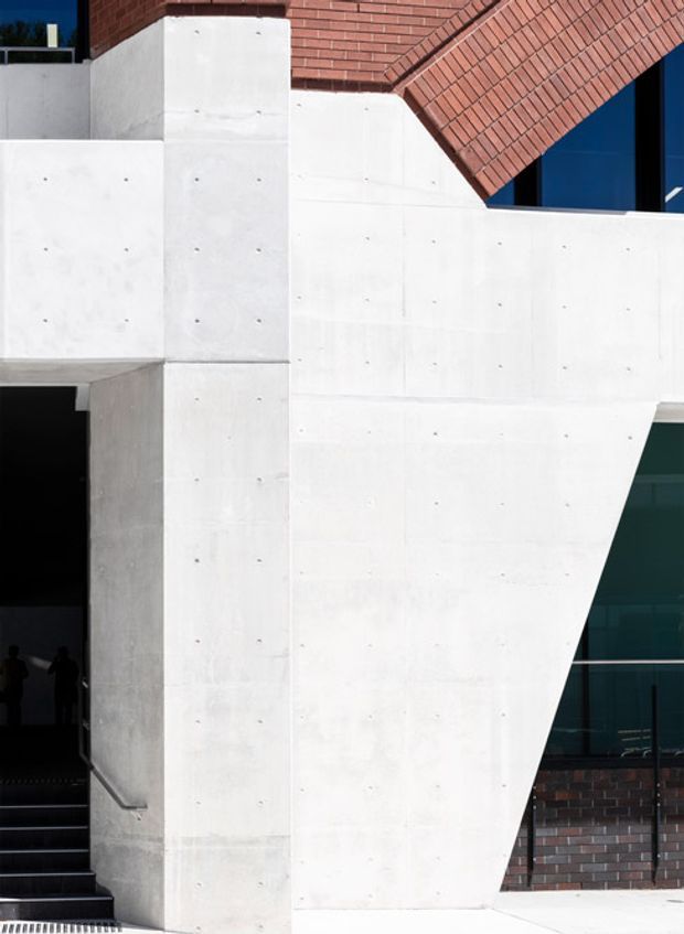 Close-up exterior architectural view dominated by sculptural white cast-concrete walls and angular projecting forms. Red brick roofline and brick wall sections contrast with dark blue-black glazing. A recessed stair entry is visible at the left, but the building facade is the primary subject. The composition is minimalist and strongly geometric, with exposed concrete panel joints and fixing marks.