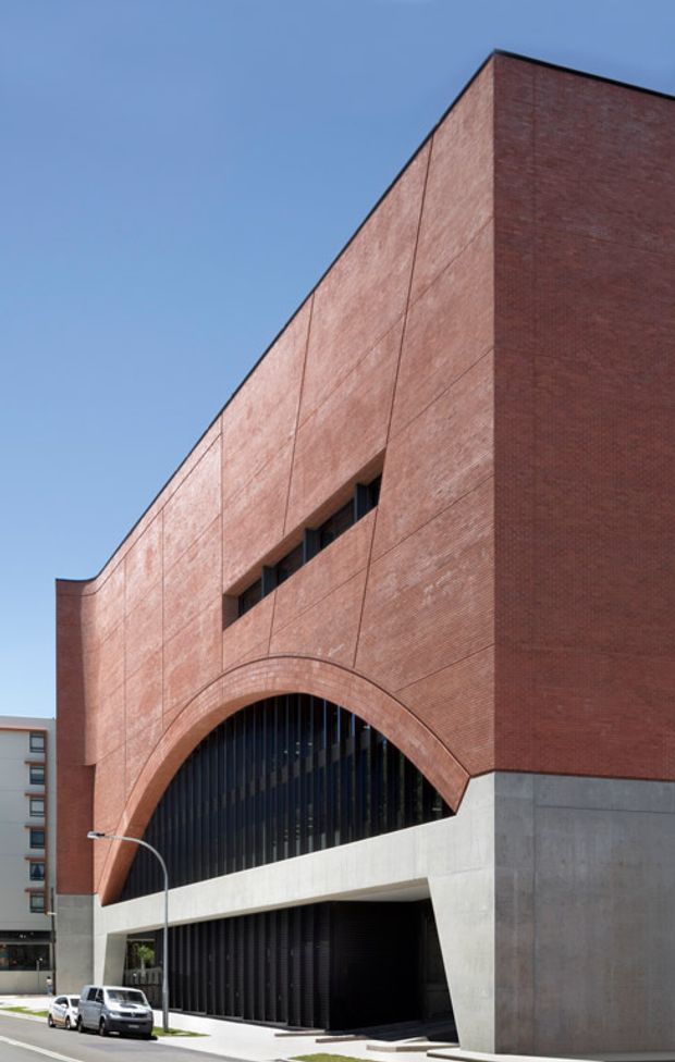A close-up exterior architectural view of a tall urban building with a monolithic red brick facade, large arched brick opening, dark vertical glazing or louvres, and a substantial exposed concrete base. The building has a contemporary geometric composition with strong verticality and minimal detailing, photographed from street level against a clear blue sky.