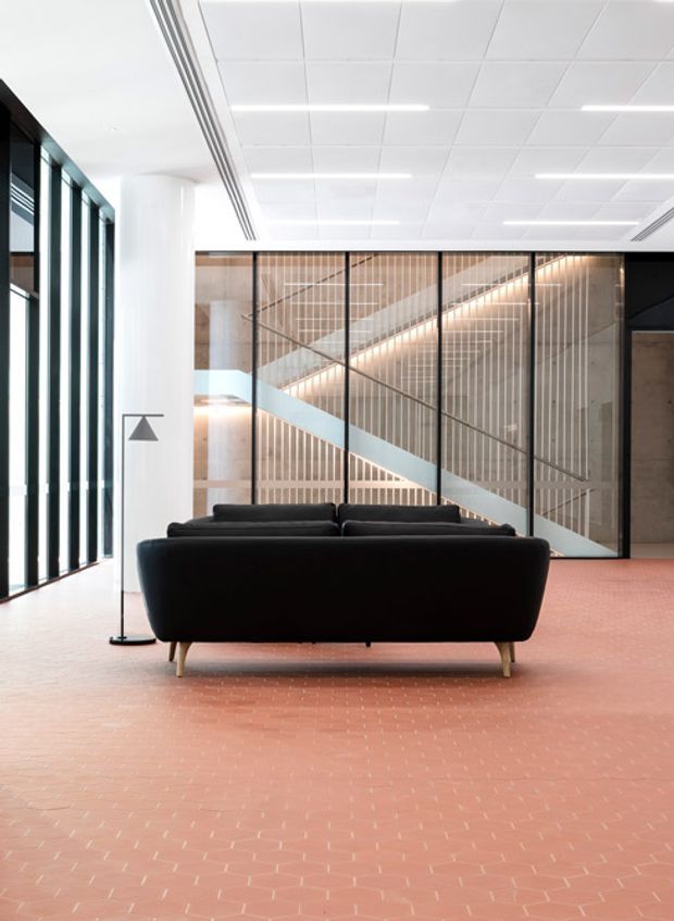 A contemporary commercial lounge or reception area with a black sofa centered on a terracotta-red tiled floor. Full-height dark-framed glazing lines the left wall, while a large black-framed glazed partition reveals a stairway behind it, featuring pale timber vertical battens and a light blue diagonal balustrade. The space has a white suspended grid ceiling with linear recessed lighting, white walls, and a minimalist floor lamp.