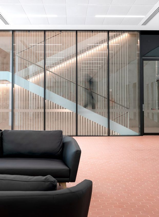 Contemporary commercial interior lounge with black leather seating in the foreground, a salmon-pink patterned tiled floor, and a glazed internal partition revealing a stairway behind it. The partition combines dark metal framing, vertical timber slats, diagonal stair geometry, and translucent diagonal privacy bands. A suspended grid ceiling and linear lighting complete the clean, modern fit-out.
