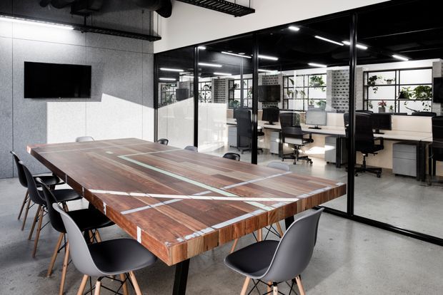 Modern commercial meeting room with a large timber boardroom table surrounded by black and grey chairs. The room has polished concrete flooring, grey textured wall panels, exposed black ceiling services, and black-framed glass partitions overlooking an open-plan office with desks, shelving, and plants.