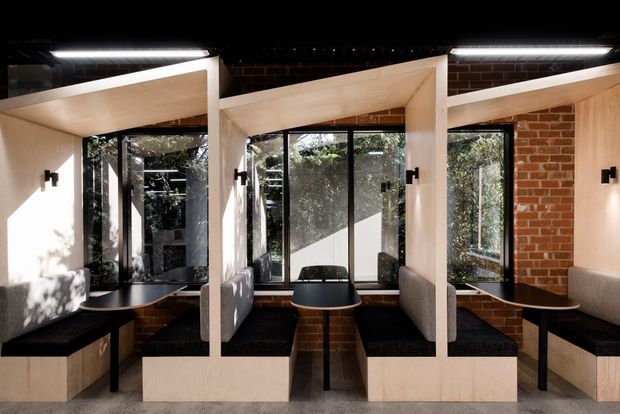 A contemporary café or hospitality interior with a series of compact booth seating bays arranged along a glazed brick wall. Pale timber or wood-finished partitions form angled overhead frames around black upholstered bench seating and small dark tables. Full-height black-framed glazing brings in abundant daylight, while exposed red brick, black ceiling elements, and wall-mounted black lights create a refined industrial character.