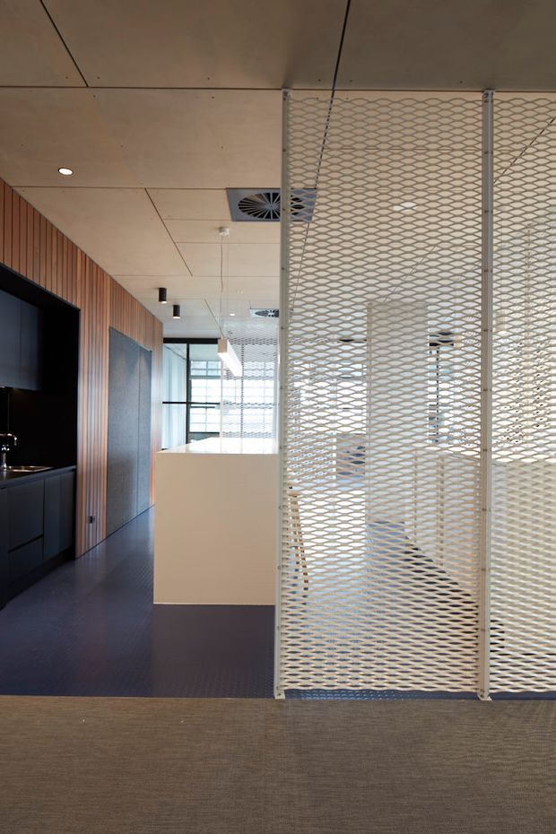 A contemporary commercial interior with an open-plan workspace screened by white perforated metal mesh partitions. The space features dark flooring, warm vertical timber wall panelling, pale timber ceiling panels, integrated dark cabinetry, recessed and surface lighting, and large glazed openings.