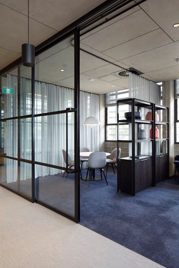 A contemporary office interior with a glazed black metal-framed meeting room enclosure, blue carpet, exposed concrete ceiling panels, sheer curtains, and a dark built-in shelving divider displaying decorative objects. A small meeting table with upholstered chairs sits inside the enclosed room, with large windows providing natural light.