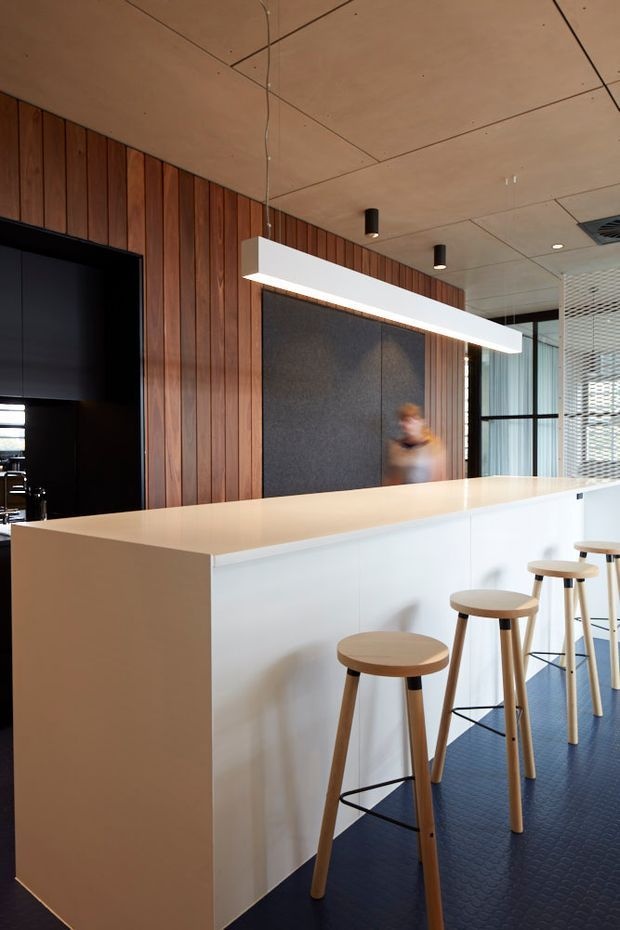 A contemporary commercial interior featuring a long white reception or service counter with timber stools, a warm vertical timber-panelled wall, dark acoustic wall panels, a suspended linear light, black-framed glazing, and a patterned dark floor. The space has a clean, refined workplace or hospitality fit-out with neutral materials and warm wood accents.