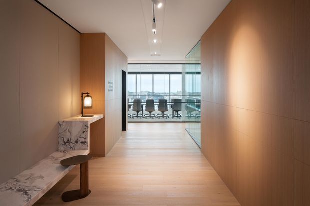 A refined contemporary office interior with a warm timber-lined corridor leading to a glazed meeting room or workspace. The space features pale timber flooring, full-height timber wall panelling, recessed and track lighting, and a sculptural marble console with a small lamp. Office chairs and desks are visible through the floor-to-ceiling glazing, with an urban view beyond.