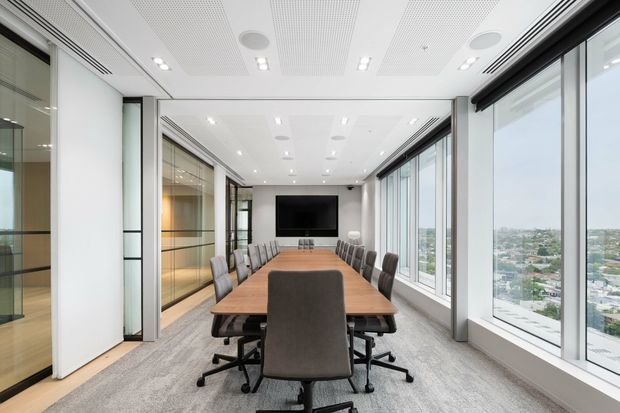 Bright contemporary boardroom with a long wood meeting table, upholstered swivel office chairs, a wall-mounted display, extensive floor-to-ceiling windows, glass-partitioned side rooms, and a perforated acoustic ceiling. The room overlooks a dense urban landscape from a high level.