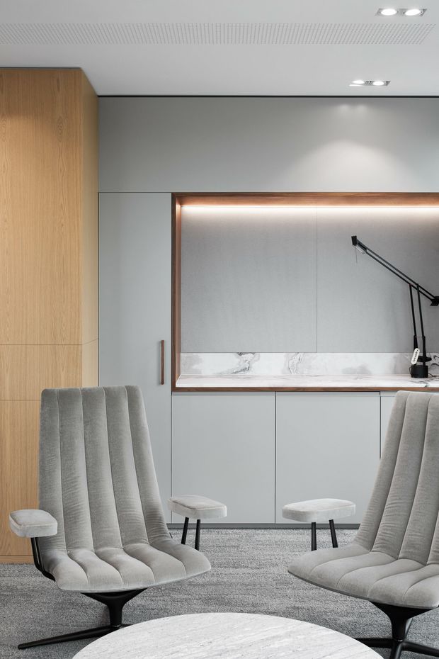 A refined contemporary office or lounge with pale grey built-in cabinetry and wall panels, a warm timber storage wall, and a recessed illuminated display niche edged in timber. Sculptural grey upholstered lounge chairs surround a round pale stone table, while a black articulated task lamp and discreet recessed ceiling lights provide focused illumination. The space has a quiet, minimalist executive feel with textured grey carpeting and a perforated acoustic ceiling.