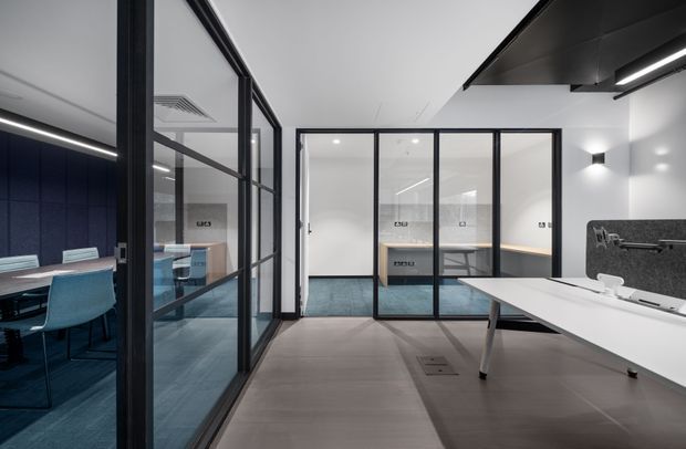Contemporary commercial office interior with glazed meeting rooms, black-framed glass partitions, open workstations, and a large white meeting table. The space uses a restrained palette of white, charcoal, black, blue carpet, and grey flooring, with linear and wall-mounted lighting.