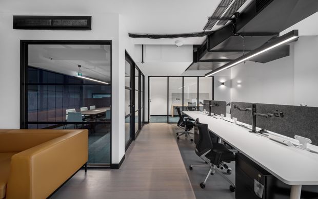 Modern open-plan commercial office interior with a long white shared workstation, multiple ergonomic black office chairs, computer monitors, and low grey acoustic desk screens. Black-framed glass partitions enclose meeting rooms, while a tan upholstered lounge seat appears in the foreground. The space has exposed dark ceiling services and linear LED lighting, with a clean monochrome palette and warm timber-look flooring.