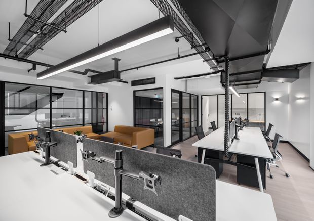 A bright modern open-plan office with rows of white workstations, computer monitor privacy screens, black-framed glass-partitioned meeting rooms, and a lounge area with tan sofas. Exposed black ceiling services, cable trays, suspended linear lighting, and visible ductwork create an industrial commercial interior.