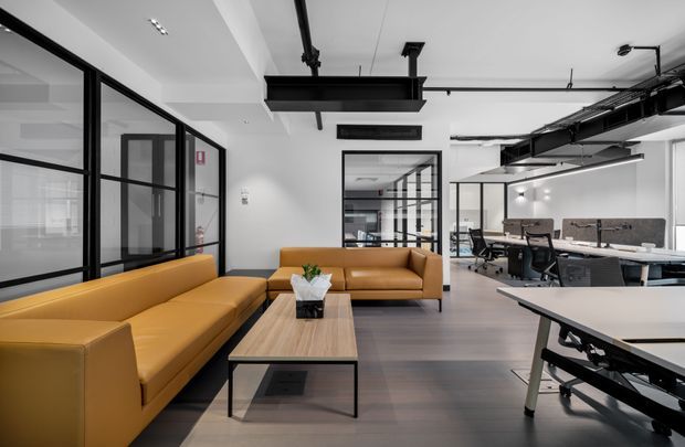 Contemporary open-plan office interior with a casual lounge area in the foreground and workstations visible beyond. Mustard-tan sectional sofas surround a light timber coffee table, while black-framed glass partitions, exposed ceiling services, suspended linear lighting, and white walls create a clean industrial workplace aesthetic.