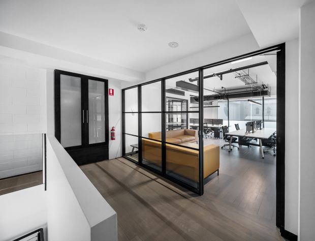A bright contemporary commercial office interior viewed from a landing or corridor. Black-framed glazed partitions enclose a meeting room with mustard upholstered seating and a table, while an open-plan workspace with desks and exposed ceiling services is visible beyond. The space has white walls and ceiling, dark timber-look flooring, and industrial-style black metal framing.
