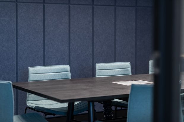 A dark, refined meeting or conference room with a large rectangular dark wood table, upholstered blue-grey office chairs, and floor-to-ceiling dark blue acoustic wall panelling. The space is partially obscured at the right edge by a foreground structural element or partition.