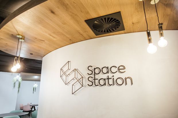 Interior view of a contemporary commercial space branded “Space Station.” A curved white partition wall features a geometric metal logo and lettering, while the ceiling combines warm timber panels with dark exposed beams. Pendant lights and a ceiling vent are visible, with a small seating/work area partially visible beyond the partition.