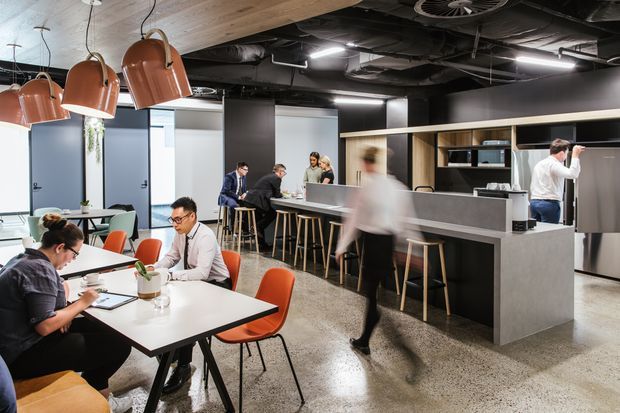 A contemporary commercial office hospitality area with shared dining and work tables, a long central bar or kitchenette with stools, black cabinetry, polished concrete flooring, exposed dark ceiling services, timber ceiling panels, and oversized copper pendant lights. Several people are working or socialising, and glazed meeting rooms are visible in the background.