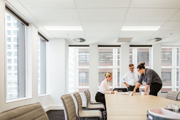 Bright contemporary commercial meeting room with a large light-wood conference table, upholstered office chairs, floor-to-ceiling windows, white walls, suspended acoustic ceiling tiles, and recessed linear lighting. Several people are gathered around the table, with an urban office building visible through the windows.