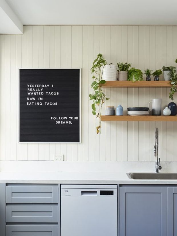 Bright, compact kitchen with pale vertical wall panelling, a white benchtop, grey-blue shaker-style cabinetry, integrated dishwasher, and stainless-steel sink tap. Two timber floating shelves display crockery, ceramics, and trailing indoor plants. A black letterboard adds a playful decorative accent.