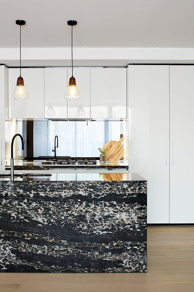 Bright contemporary kitchen featuring a large waterfall island clad in dramatic dark natural stone with dense white and warm veining. The space has glossy white floor-to-ceiling cabinetry, integrated appliances, black tapware and fixtures, timber flooring, mirrored splashback surfaces, and two pendant lights with glass shades. The composition is clean-lined and high-contrast, with abundant daylight.