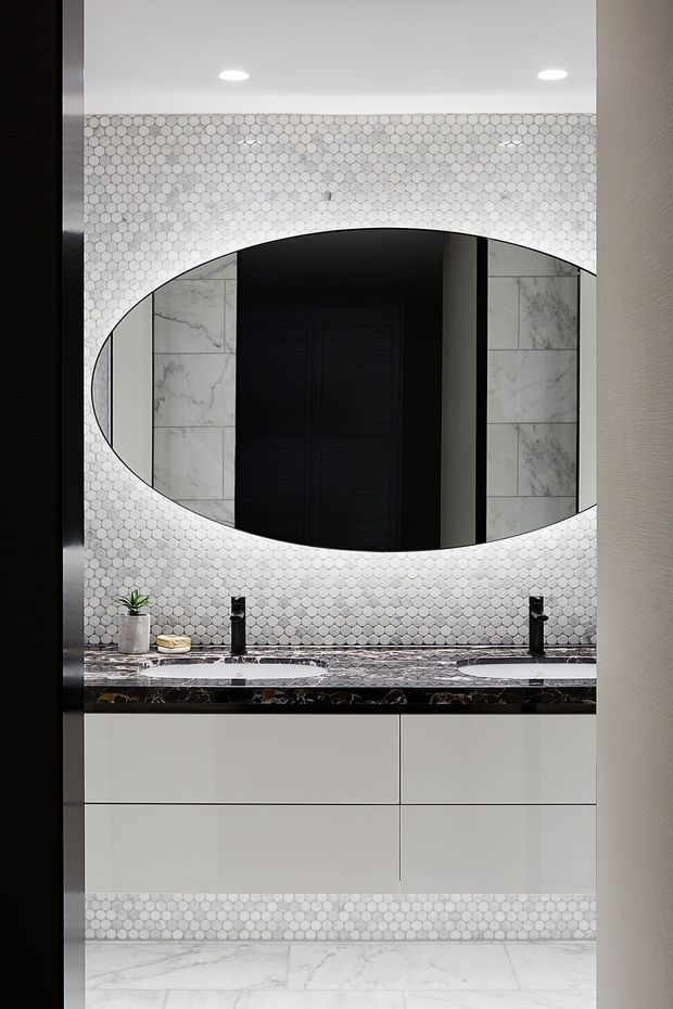 A refined contemporary bathroom vanity viewed through a doorway, featuring a large oval mirror, white hexagonal mosaic wall tiles, marble-look wall and floor tiles, a dark veined stone benchtop, twin black tapware and basins, and streamlined white handleless cabinetry. The mirror reflects a dark interior doorway and tiled side walls.