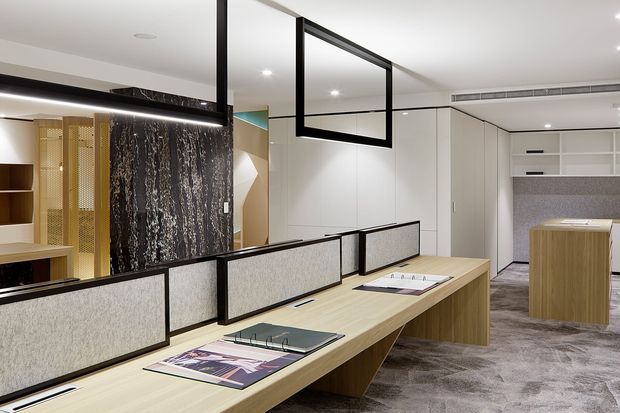 Contemporary office or commercial interior with long light-wood workstations, low black-framed acoustic privacy screens, integrated cabinetry, recessed lighting, and grey carpet. A dark textured feature wall and gold-toned perforated screen add visual interest, while a kitchenette or service counter is visible at the rear.