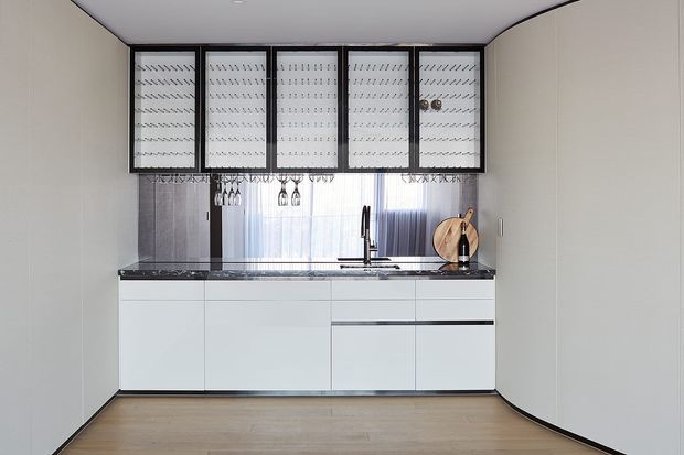 A compact contemporary kitchen with a full wall of white handleless base cabinetry, a dark stone benchtop, and a stainless-steel sink and tap. Black-framed upper glass-front cabinets feature patterned translucent panels, with hanging stemware beneath. A softly veiled window provides natural light, while timber boards and pale walls create a clean, restrained palette.