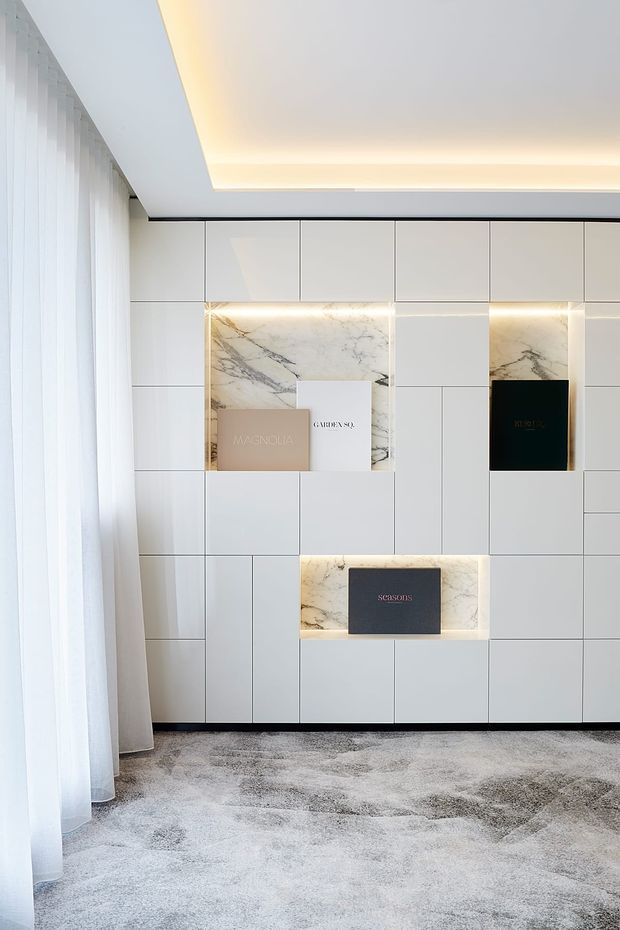 A bright, refined interior featuring a full-height white built-in display wall with a grid of flush panels and several recessed illuminated niches. The niches are lined with veined white marble and contain sample books or presentation boxes. Sheer white curtains cover a full-height window along the left side, while a pale textured carpet and concealed cove lighting create a soft, minimalist atmosphere.