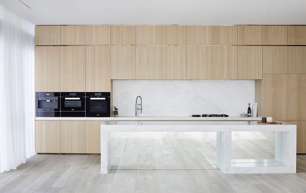 A bright, minimalist contemporary kitchen with a full wall of light timber cabinetry, integrated black appliances, a pale marble-look splashback, and a large white waterfall island with open display shelving. Light timber flooring and sheer curtains create an airy, refined interior.
