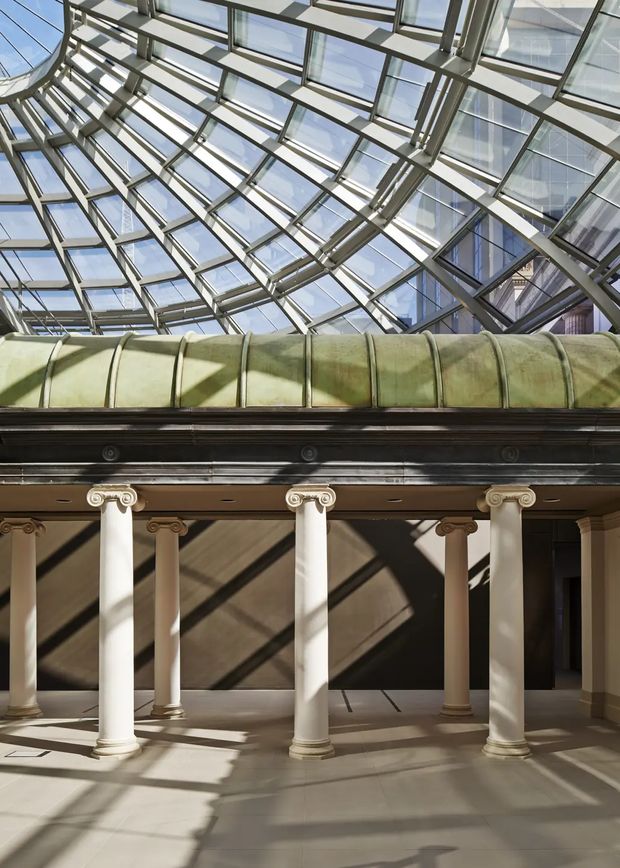 A grand interior atrium beneath a large glazed skylight and steel-framed glass roof. Classical white columns with ornate capitals support a dark cornice and green barrel-vaulted canopy, while strong sunlight casts geometric shadows across the pale stone floor and walls.