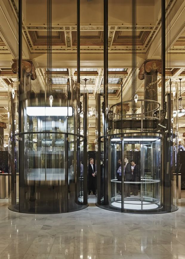 Grand ornate commercial interior lobby featuring mirrored glass elevator shafts, dark metal framing, polished light stone flooring, marble-clad columns, and an elaborate coffered ceiling with geometric decorative motifs and skylight-like panels. The space has a luxurious historic character with contemporary glass elevators and dramatic vertical lines.