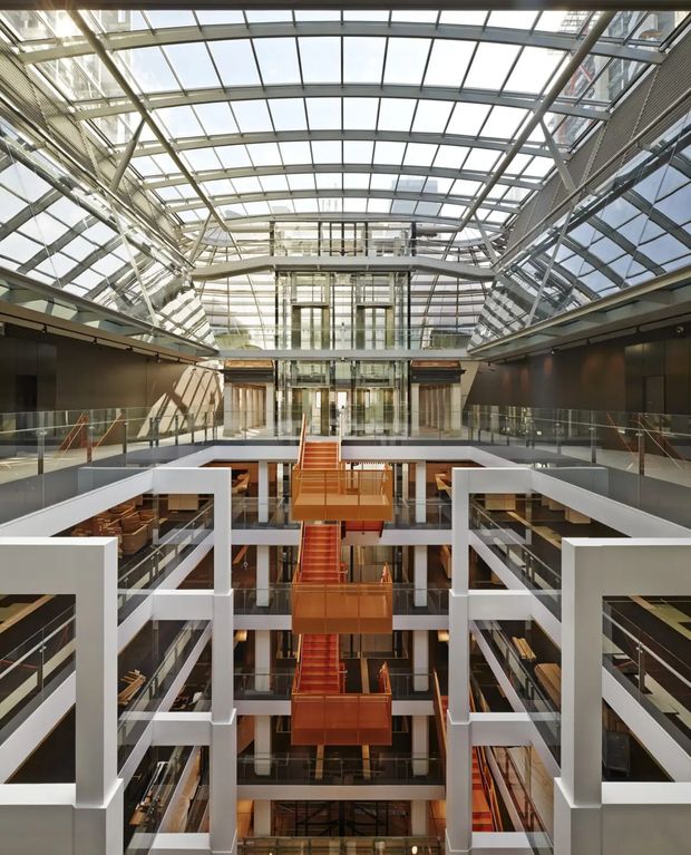 A dramatic interior view of a large multi-level commercial atrium, likely a library, institutional building, or office complex. White structural balconies and bridges frame a central orange stairway and lift core, with extensive glazing, exposed steel framing, and a translucent vaulted roof flooding the space with daylight. The image emphasizes the building's vertical circulation, atrium void, and contemporary architectural structure.