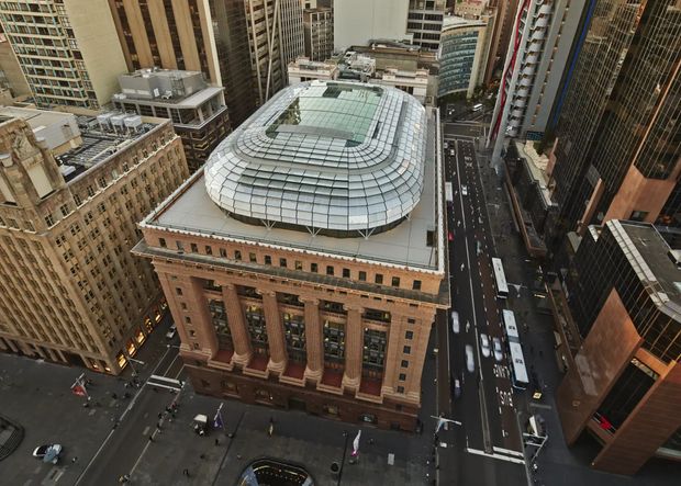 Aerial urban view of a large historic masonry civic or commercial building with monumental columns and ornate classical detailing, topped by a contemporary rounded glass-and-metal roof structure. Dense high-rise buildings and surrounding city streets are visible around it.