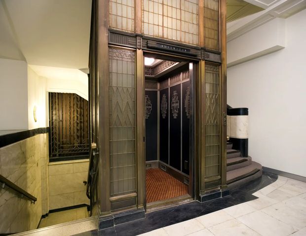 Interior view of an ornate vintage elevator lobby in an Art Deco-style building. A bronze-toned, glazed elevator surround with geometric metal detailing frames an open elevator car with dark decorative panels and a patterned floor. Marble or stone wall and floor finishes, a stair descending to the left, and a curved stair or landing to the right reinforce the refined historic commercial interior.