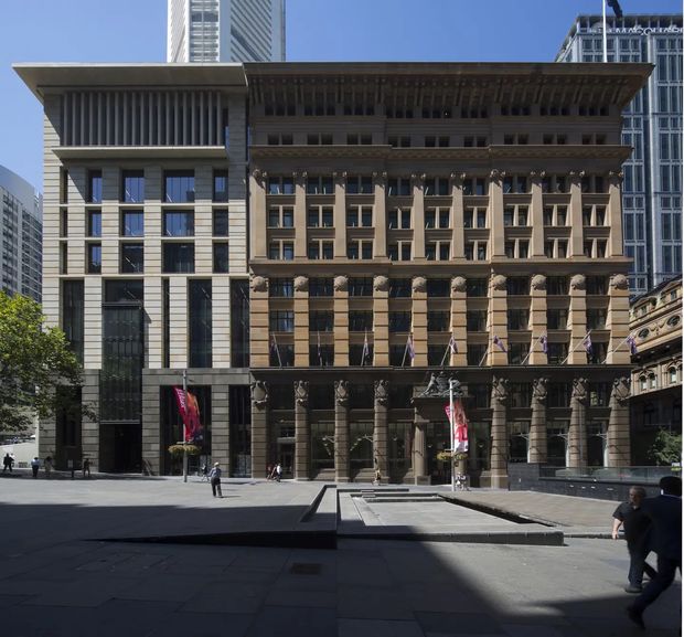 Exterior view of a prominent urban commercial building ensemble in a paved civic plaza. The main historic-looking stone facade has strong classical detailing, including repeated columns, projecting cornices, decorative capitals, recessed window bays, and a large overhanging roof-like canopy. A more contemporary stone-and-glass volume adjoins it on the left, with tall vertical fins and grid-patterned windows. High-rise buildings are visible in the background, and pedestrians occupy the plaza.