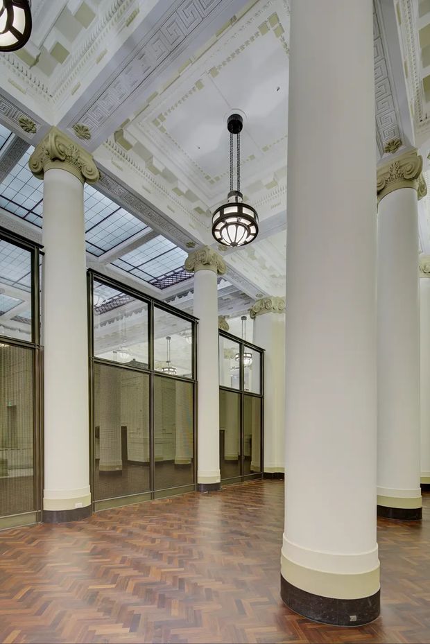 A grand historic interior hall with soaring white decorative ceilings, ornate classical cornices, large fluted columns with elaborate capitals, black-framed glazed partitions, and a suspended spherical lantern. The floor is finished in a warm herringbone timber pattern, while a large glazed roof or skylight structure brings light into the space. The architecture combines monumental classical detailing with dark metal and glass partitioning.