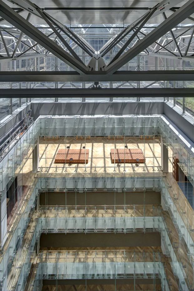Vertical view through a large multi-level interior atrium, showing extensive transparent glass balustrades and bridges, exposed steel structural trusses, and a glazed roof overhead. The space appears institutional or commercial, with several floors and a bright central void.