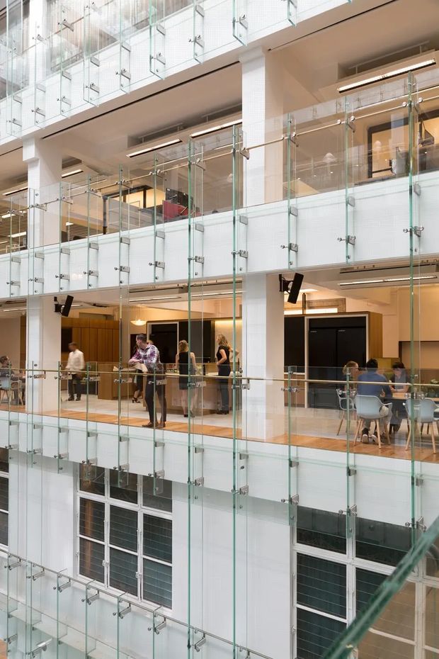 A bright multi-level commercial atrium with open circulation balconies, extensive clear glass balustrades supported by metal fittings, white structural columns, timber-toned floors, and warm wood-panelled areas. Several people are visible using the shared interior space, with dining or breakout seating on the lower level. The architecture is clean, contemporary, and strongly defined by the transparent layered walkways and tall atrium volume.