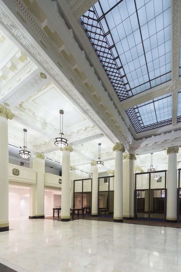 A grand, highly ornamented interior atrium or lobby with tall classical columns, elaborate white decorative plasterwork, geometric friezes, gilded capitals, suspended pendant lights, and large glazed skylights. The polished pale stone floor and black-framed glazed partitions create a bright, monumental heritage-style public space.