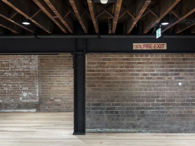 Interior architectural view of a converted industrial-style space, showing exposed timber roof joists and rafters, a black steel structural column and beam, weathered exposed brick walls, and pale timber flooring. Fire-exit signage and emergency lighting are visible, reinforcing the utilitarian commercial or adaptive-reuse character.